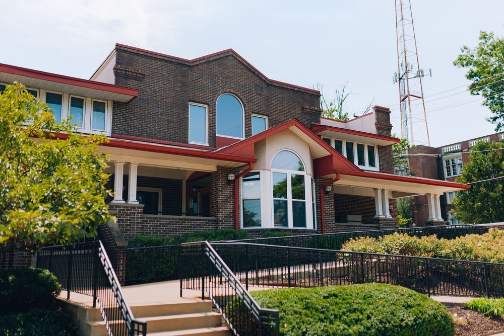 Brick building with red roof, arched windows, black railings, and stairs surrounded by lush green trees on a sunny day.