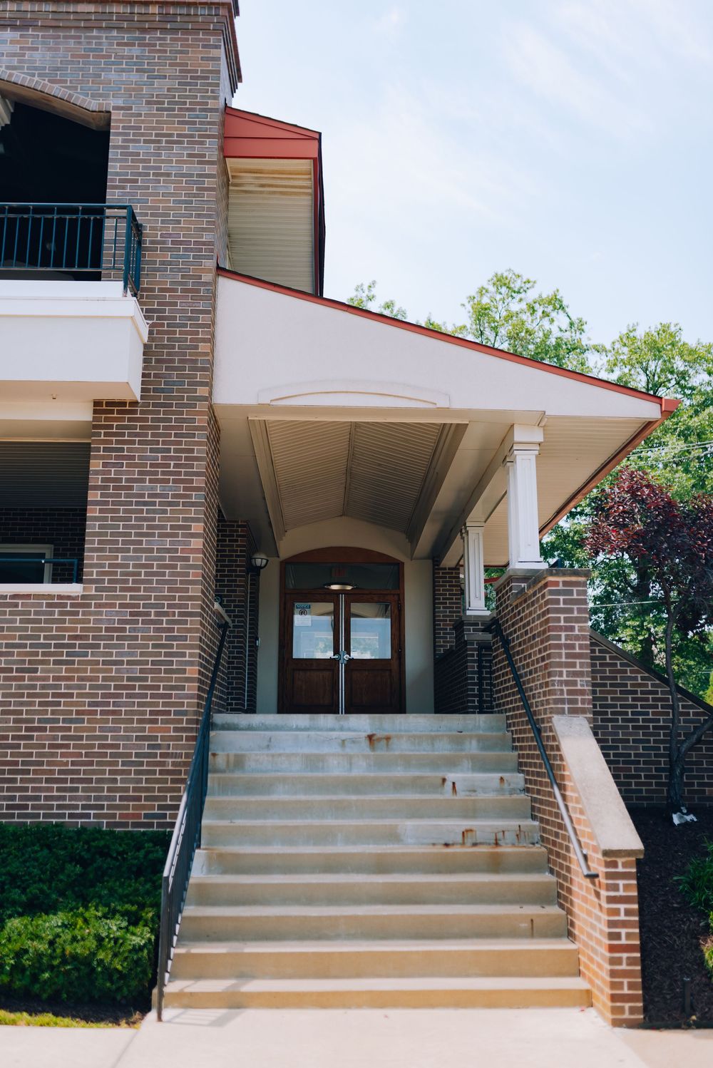 Brick building entrance with concrete steps, white columns, arched doorway, and covered portico under a partly cloudy sky.