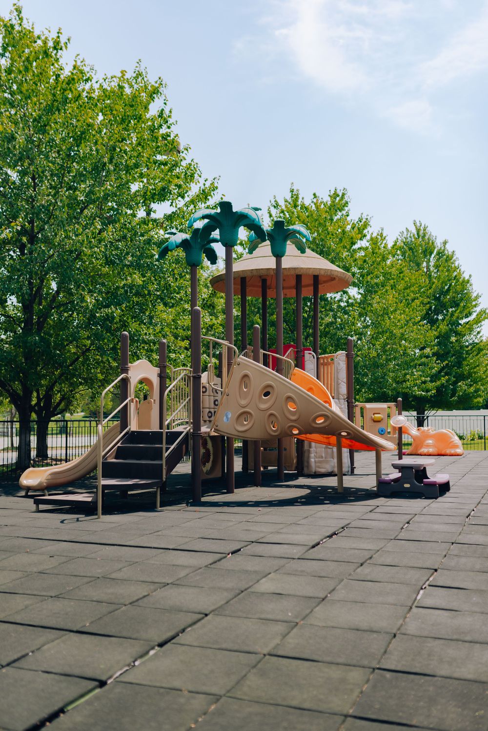 Colorful children's playground with slides and palm tree decorations surrounded by lush green trees on a sunny day.
