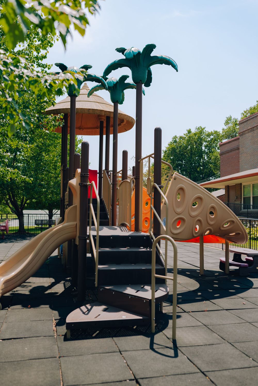 Colorful playground with palm tree decorations, slide, climbing wall, and stairs on rubber safety tiles near a school building.