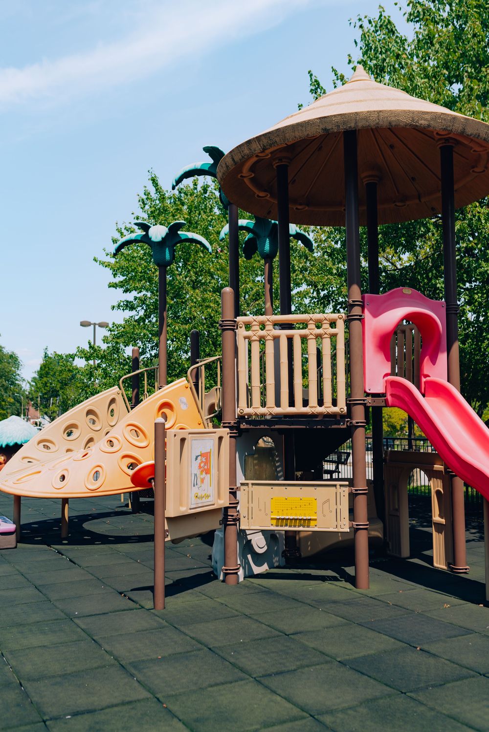 Colorful children's playground with red slide, climbing wall, and gazebo roof surrounded by green trees on rubber flooring.