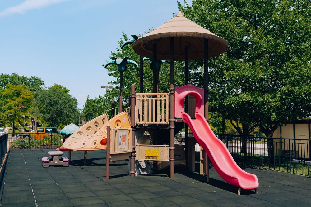 Colorful children's playground with pink slide, climbing wall, and wooden structure surrounded by green trees on rubber flooring.