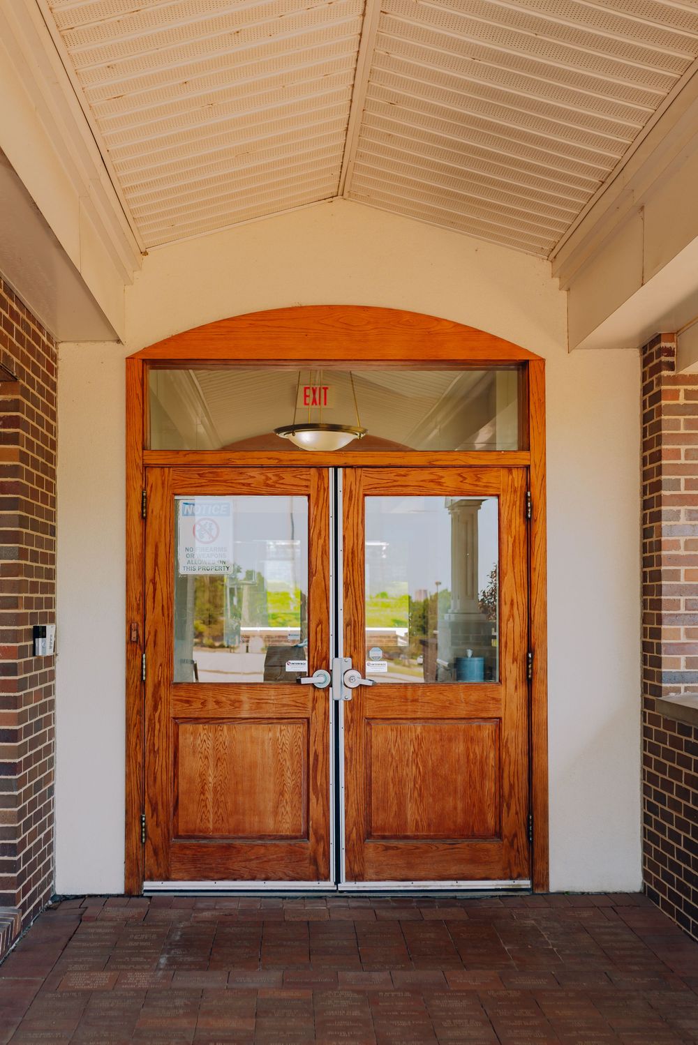 Wooden double doors with glass panels and an exit sign above, set in a brick building entrance with a white ceiling.