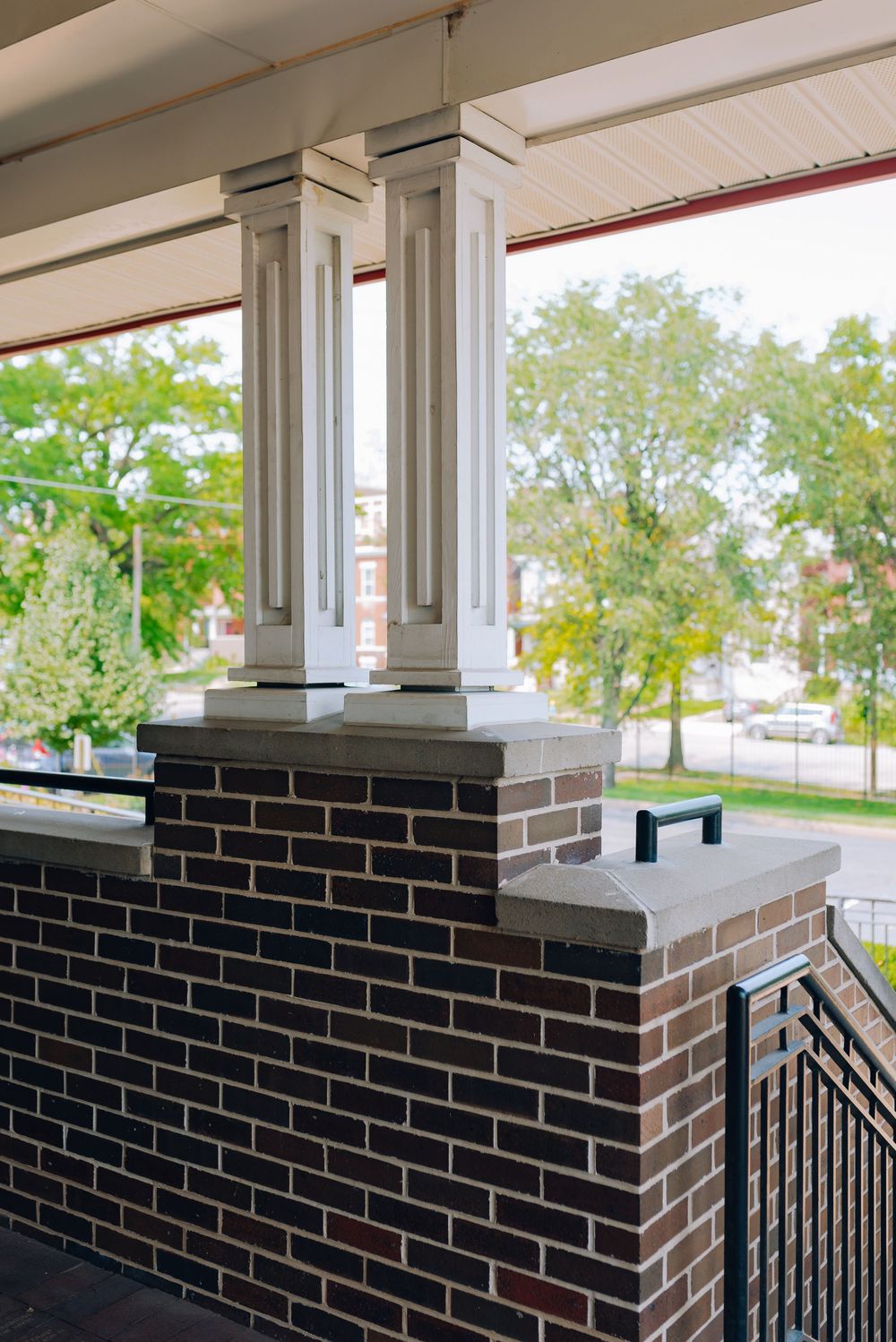 Brick porch columns with white decorative pillars, black metal railing, and lush green trees visible in the background.
