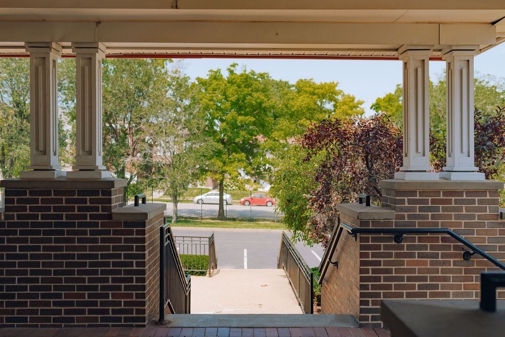 Covered brick porch with white columns overlooking a tree-lined street on a sunny day.