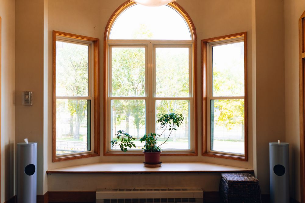Elegant wooden arched window with three panels, a potted plant on the sill, overlooking lush green trees in natural light.