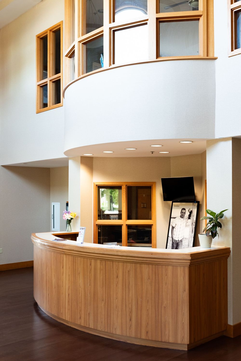 Modern medical office reception desk with curved wood paneling, TV monitor, and two-story atrium with decorative windows.