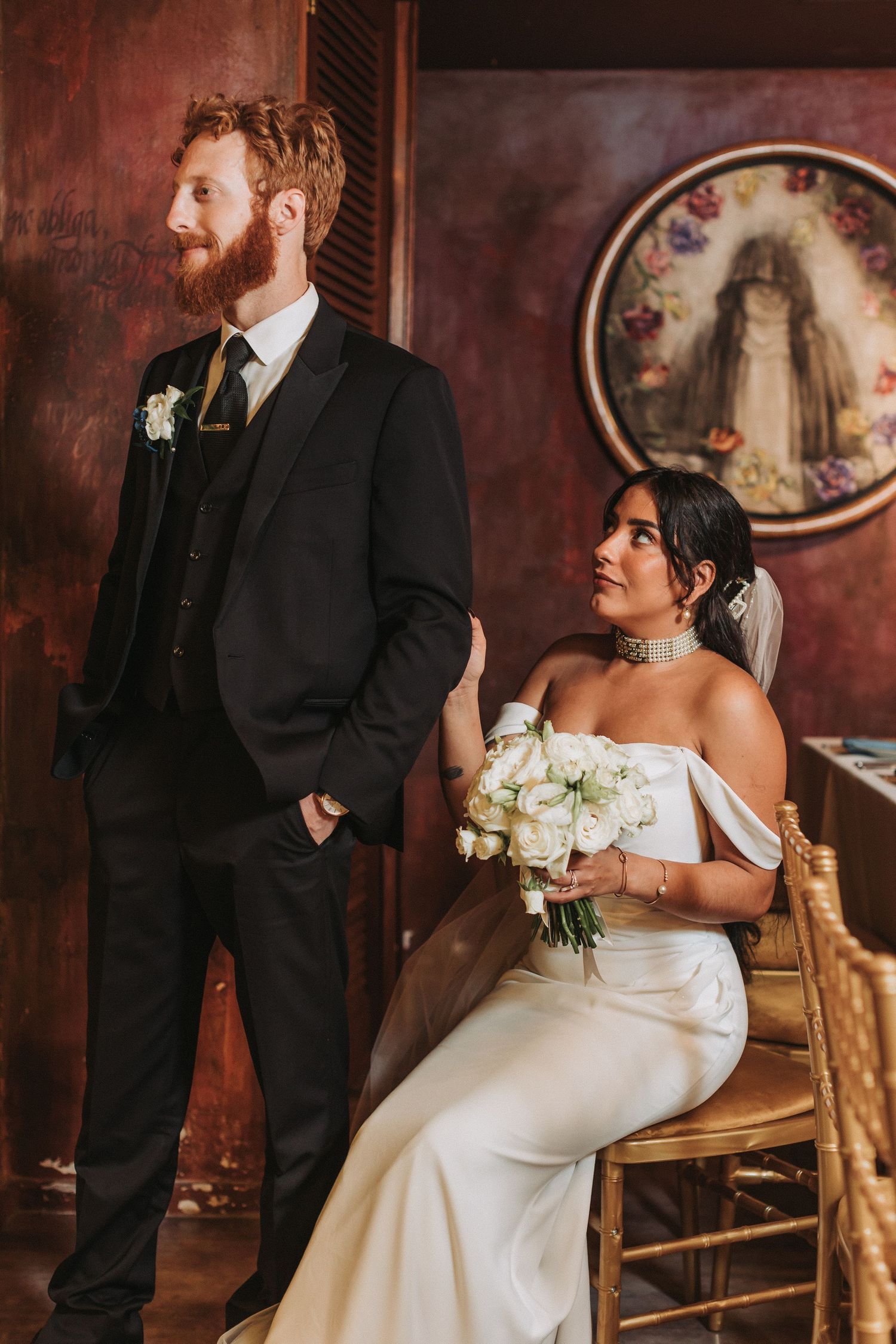 An elegantly dressed couple poses in front of a decorative circular wall piece at their wedding.
