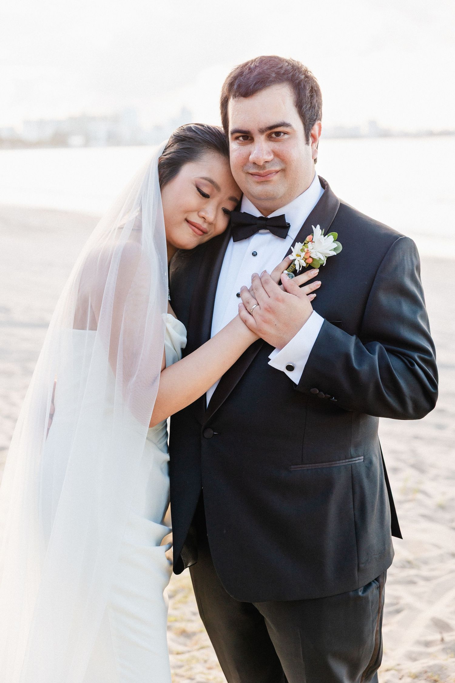 Bride in white gown and veil leans on groom in black tuxedo with boutonniere on a sandy beach.