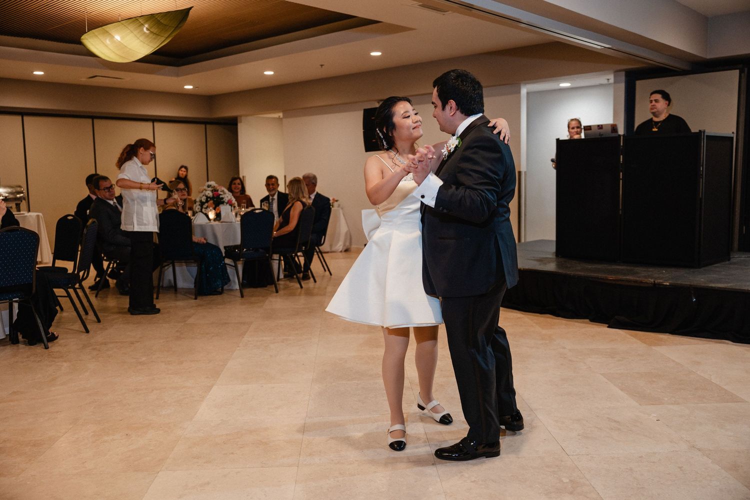 Bride and groom share their first dance at wedding reception in elegant ballroom with guests seated at tables.