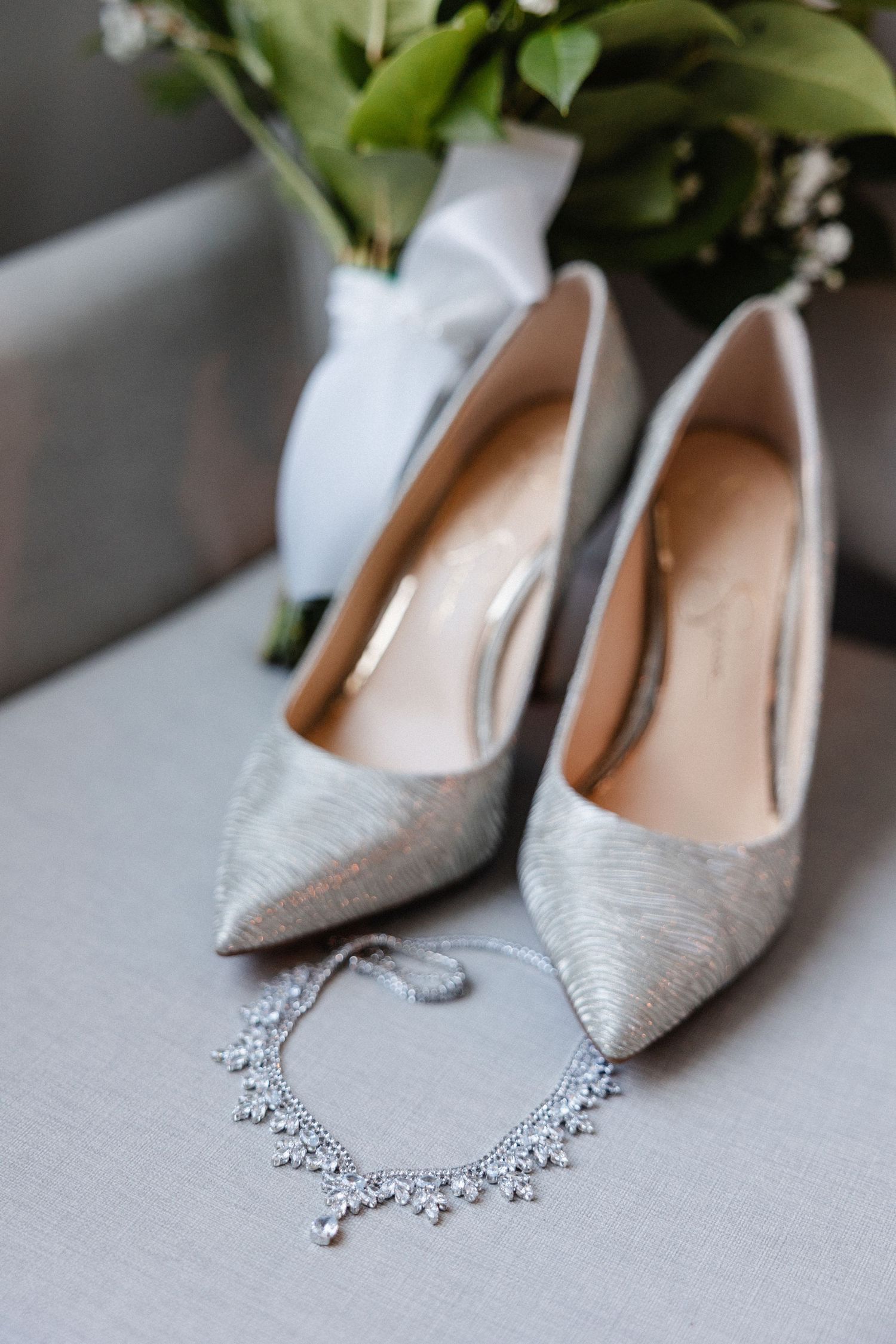 Silver glitter pointed-toe wedding heels with crystal anklet necklace and white bridal bouquet on gray surface.