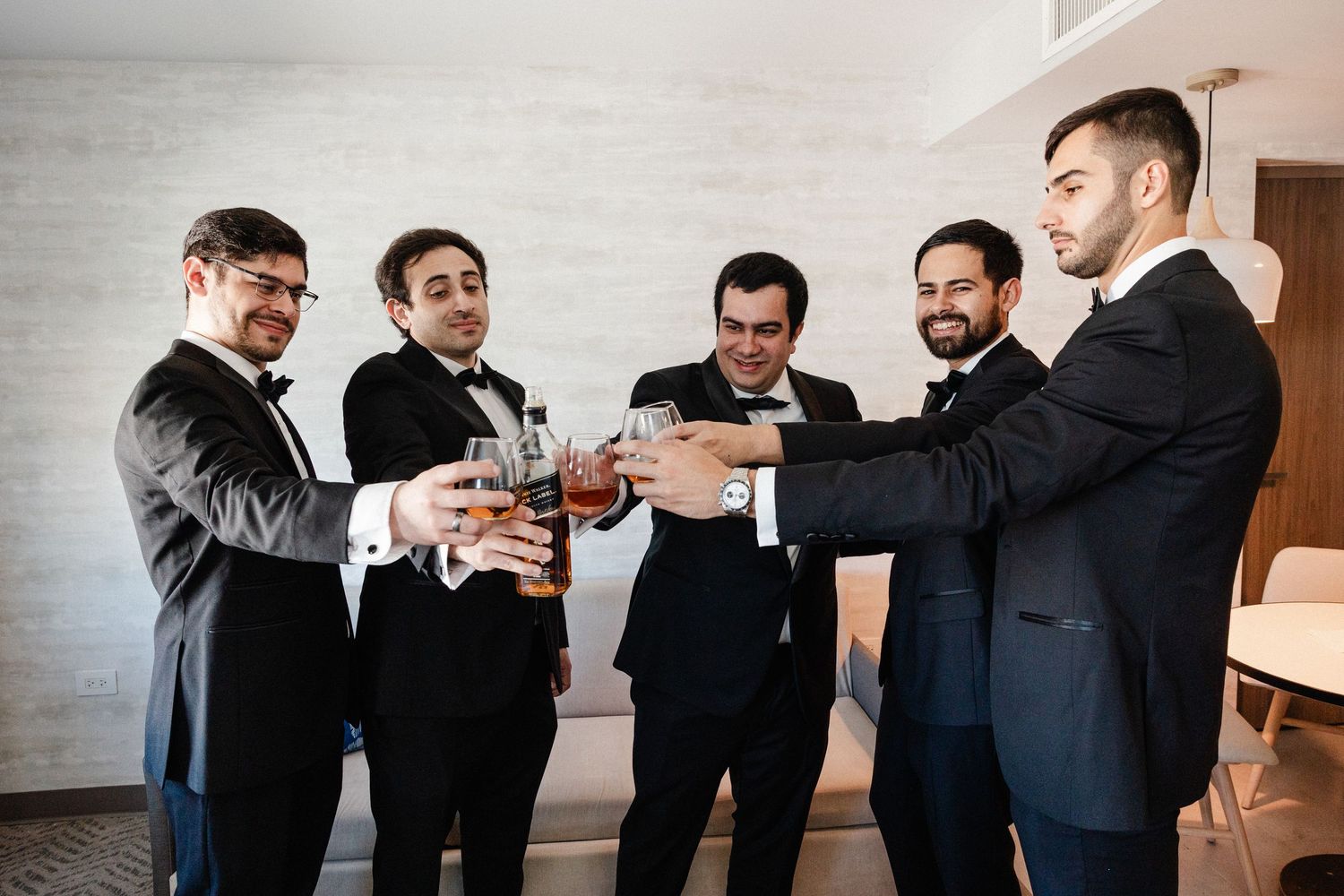 Five groomsmen in black tuxedos raising glasses in a celebratory toast before a wedding ceremony.