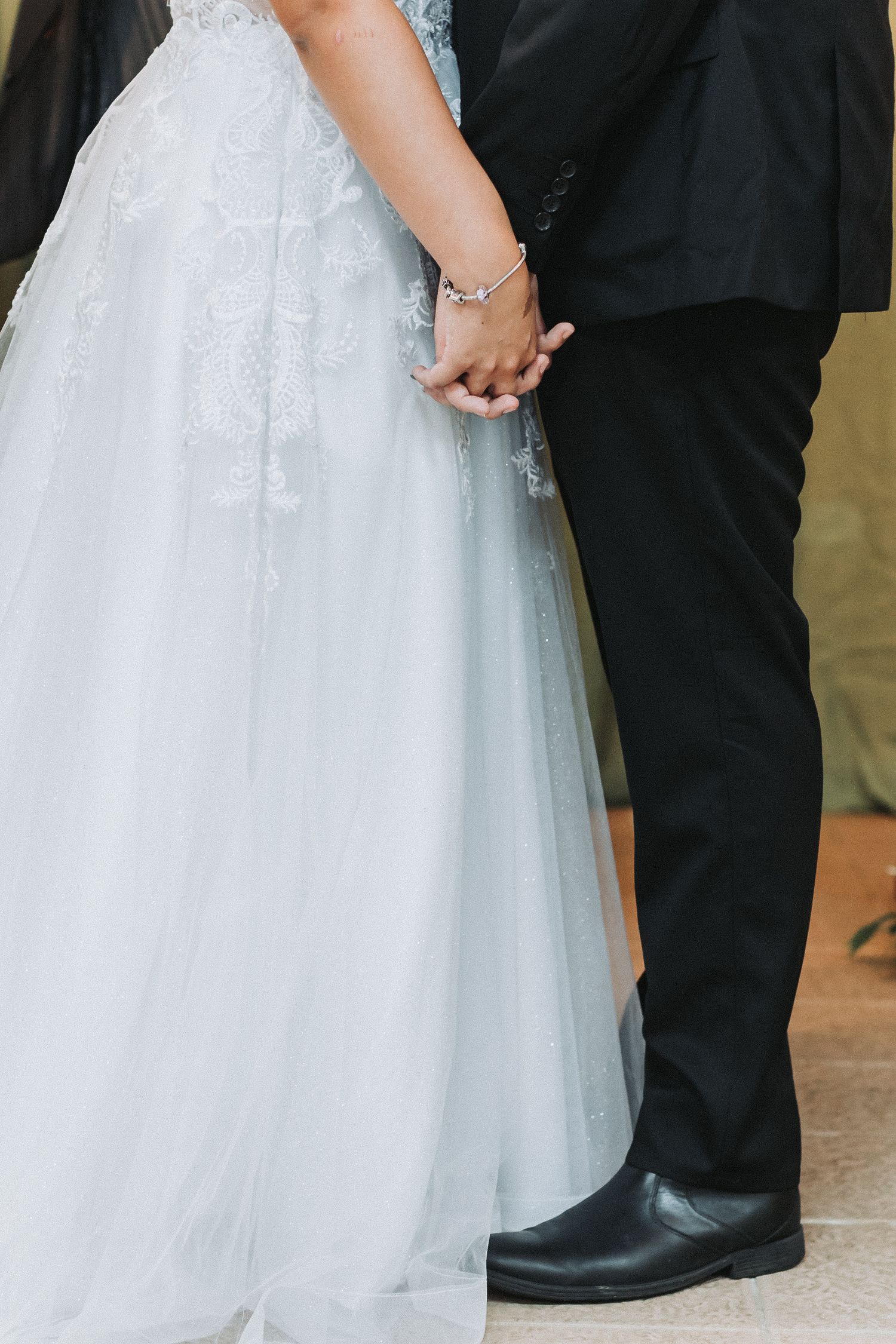 A close-up of a bride and groom holding hands, showing wedding dress details.
