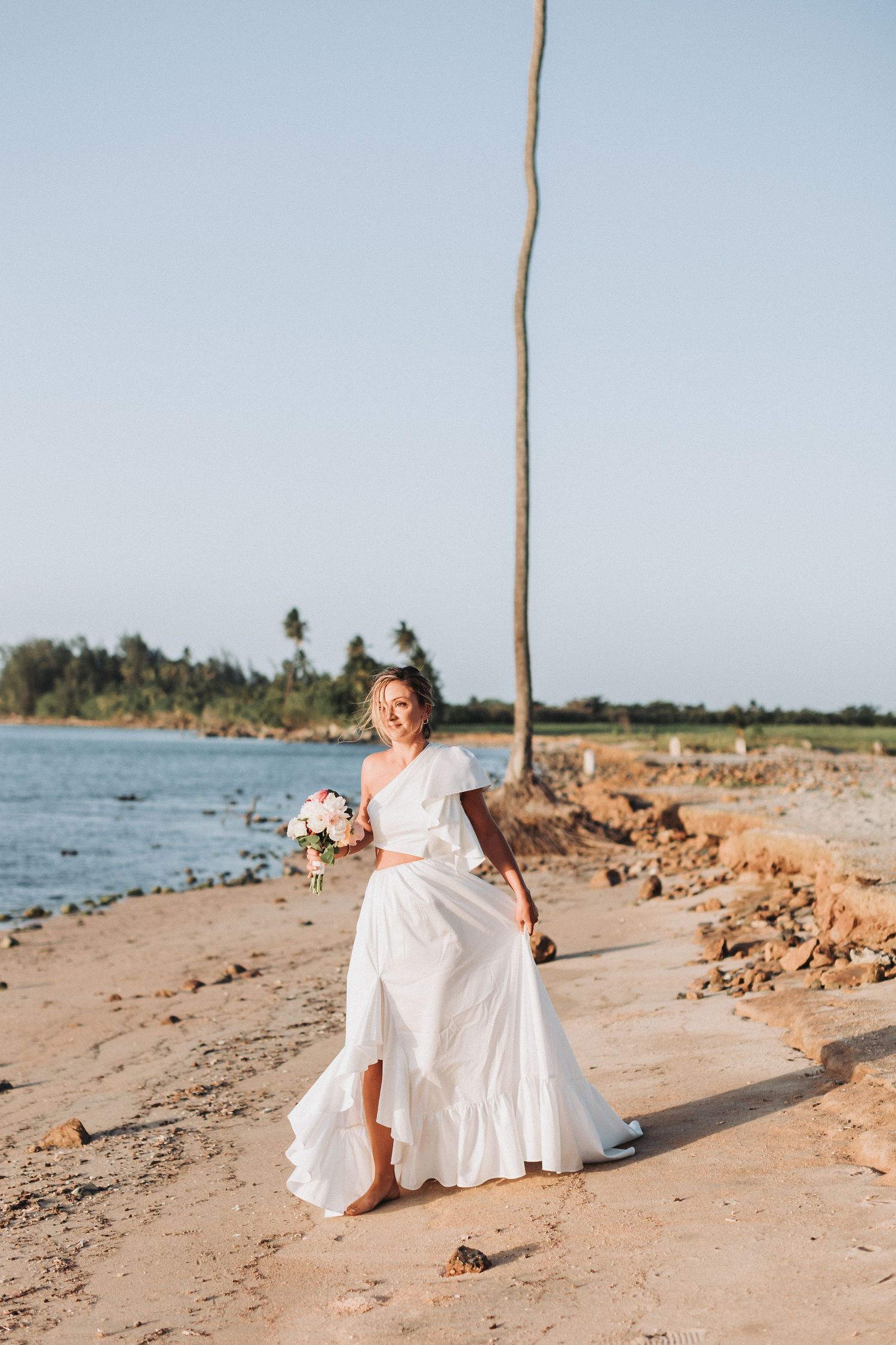 A person in a flowing white dress holds a bouquet while standing on a tropical beach.