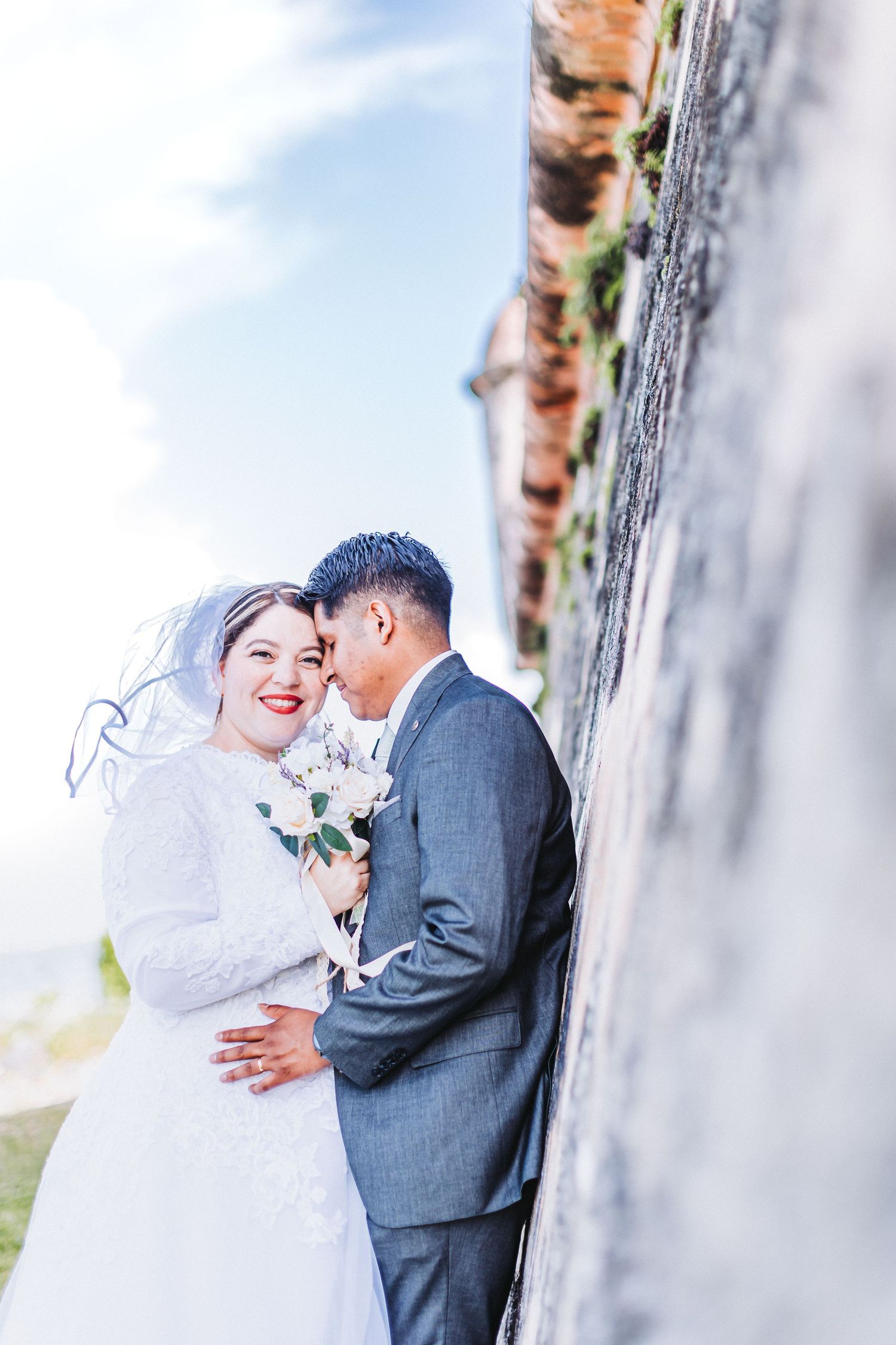 A romantic wedding photo against a weathered stone wall shows a couple embracing in white dress and grey suit.