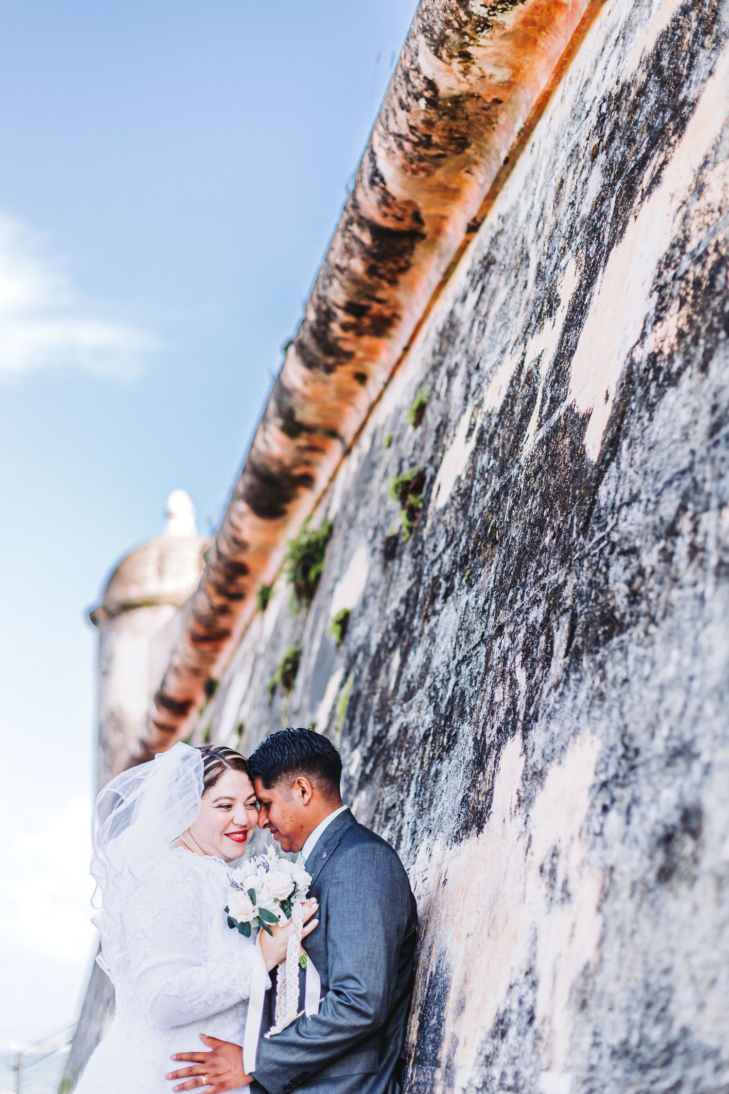 Newlyweds share an intimate moment against a textured historic wall under bright blue skies.