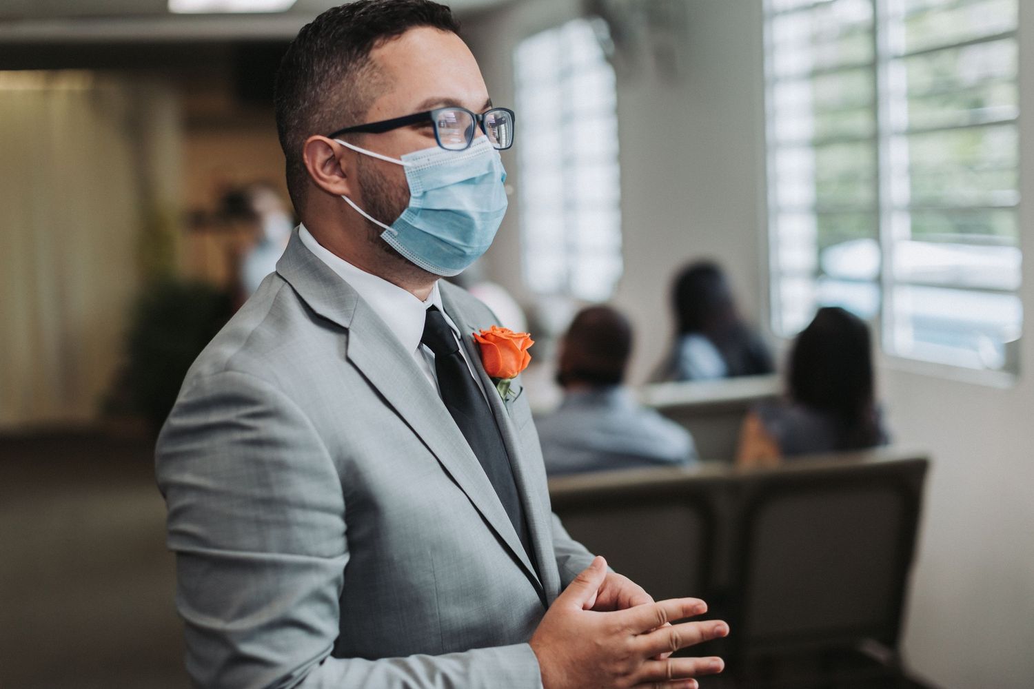 Groom in grey suit and face mask with orange boutonniere in indoor setting.