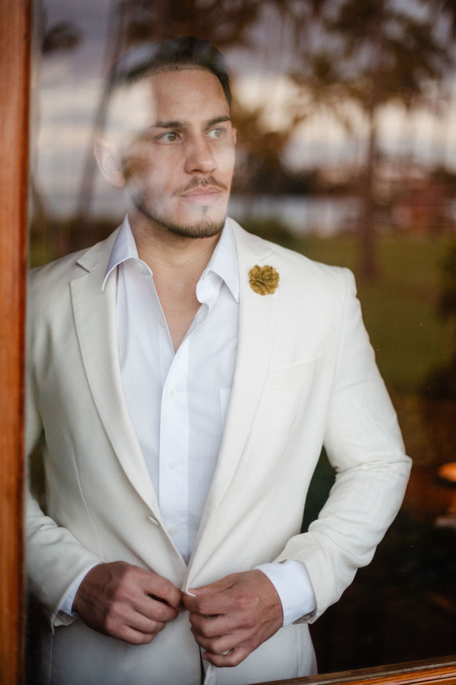 A stylish groom in a cream white suit with gold boutonniere buttoning his jacket while gazing through a window.