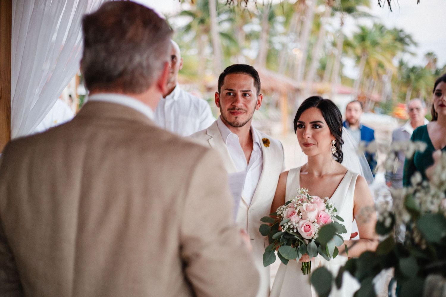 Bride and groom exchange vows at an outdoor beach wedding ceremony, bride holding a bouquet of pink roses.