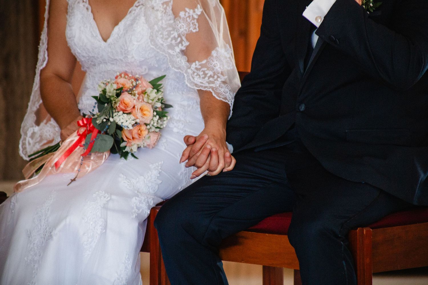 Bride in lace wedding dress holding peach bouquet sits beside groom in dark suit, holding hands at wedding ceremony.