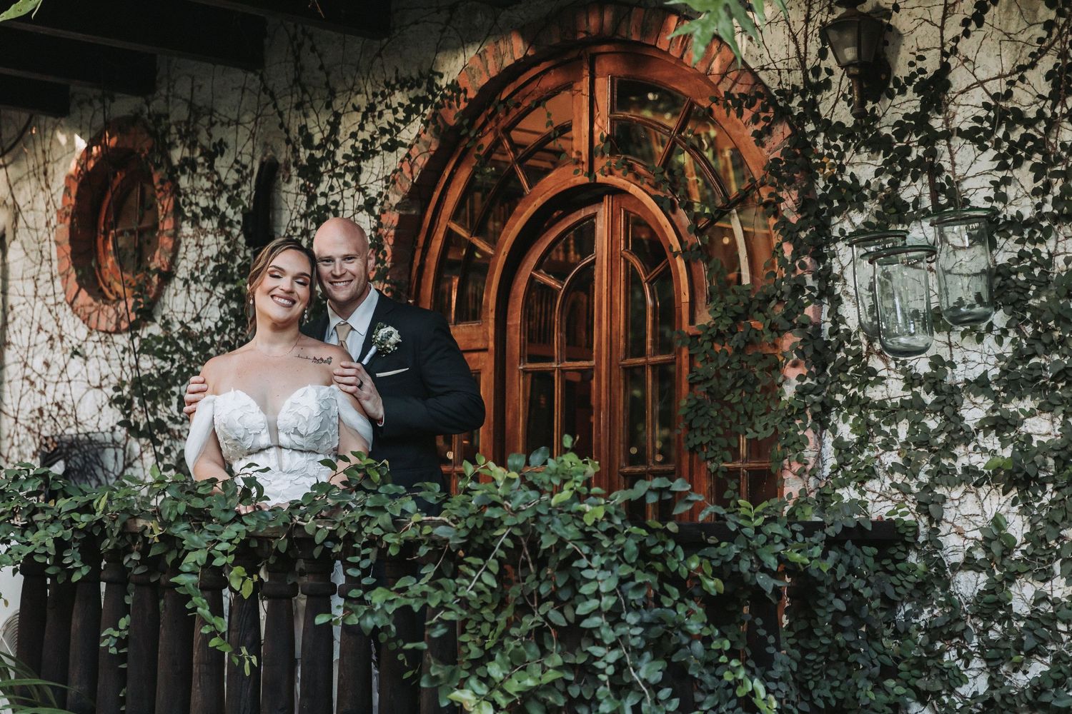 A couple stands on a balcony framed by ivy and arched wooden windows.