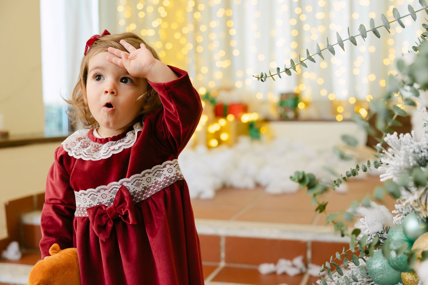 Two joyful girls laugh and play in artificial snow surrounded by Christmas gifts and festive decorations indoors.