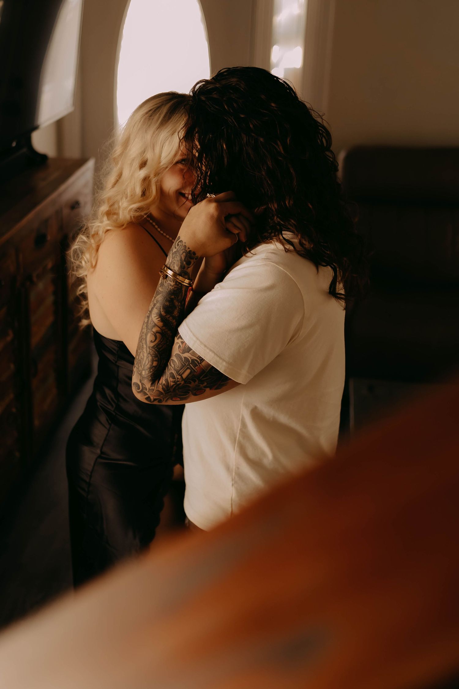 A tattooed couple shares an intimate embrace in warm moody lighting, wearing black dress and white tee.