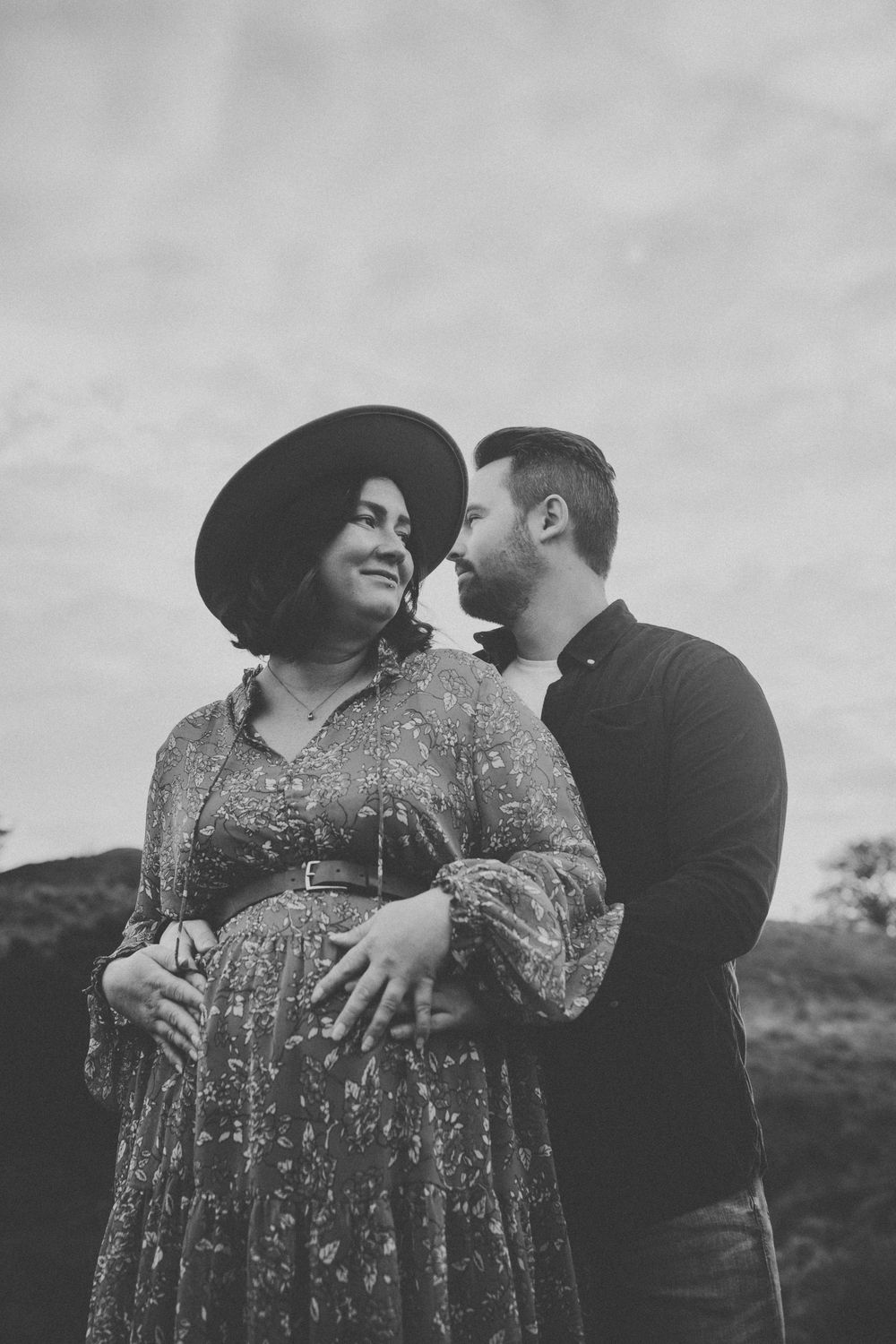 Black and white photo of a smiling pregnant woman in a floral dress and hat with her partner outdoors.