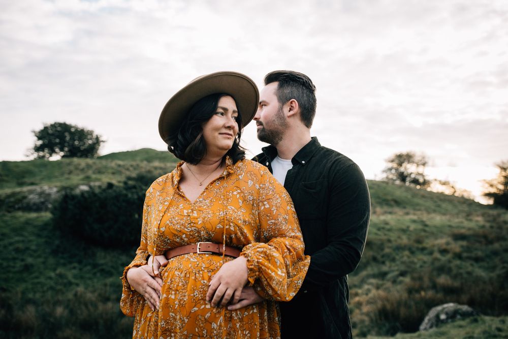 Pregnant woman in yellow floral dress and hat stands with partner in green field at dusk.