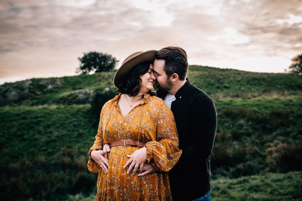 Couple sharing a tender moment outdoors, woman in yellow floral maternity dress and brown hat, man in black sweater.