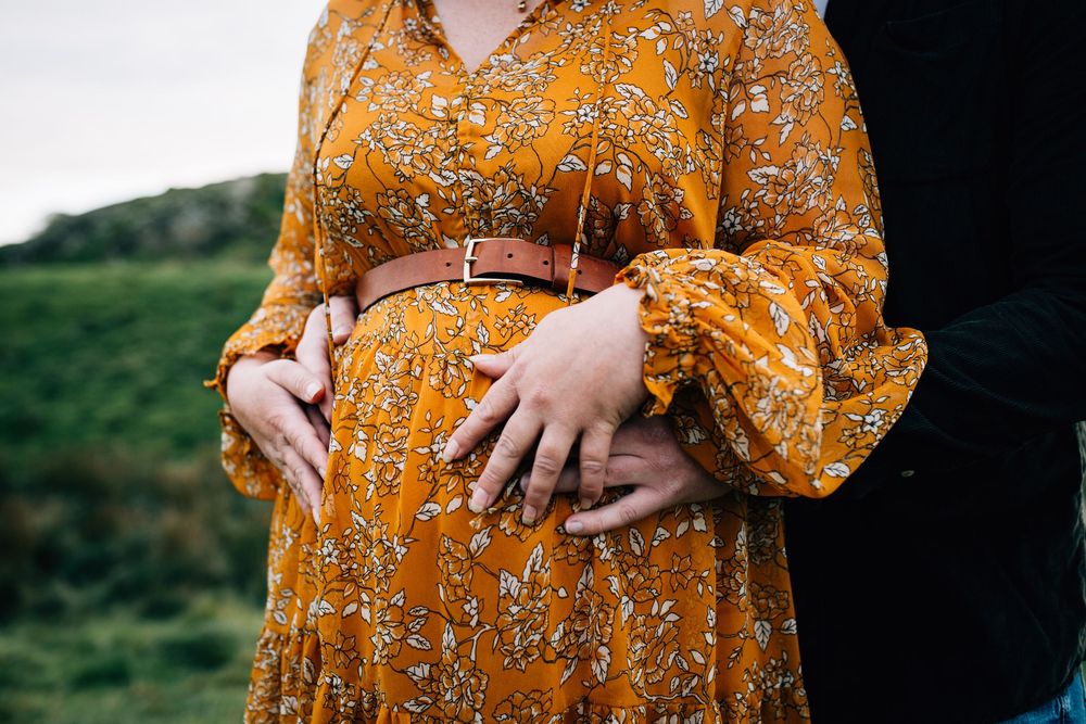 Pregnant woman in orange floral dress with belt, partner's hands cradling baby bump outdoors.