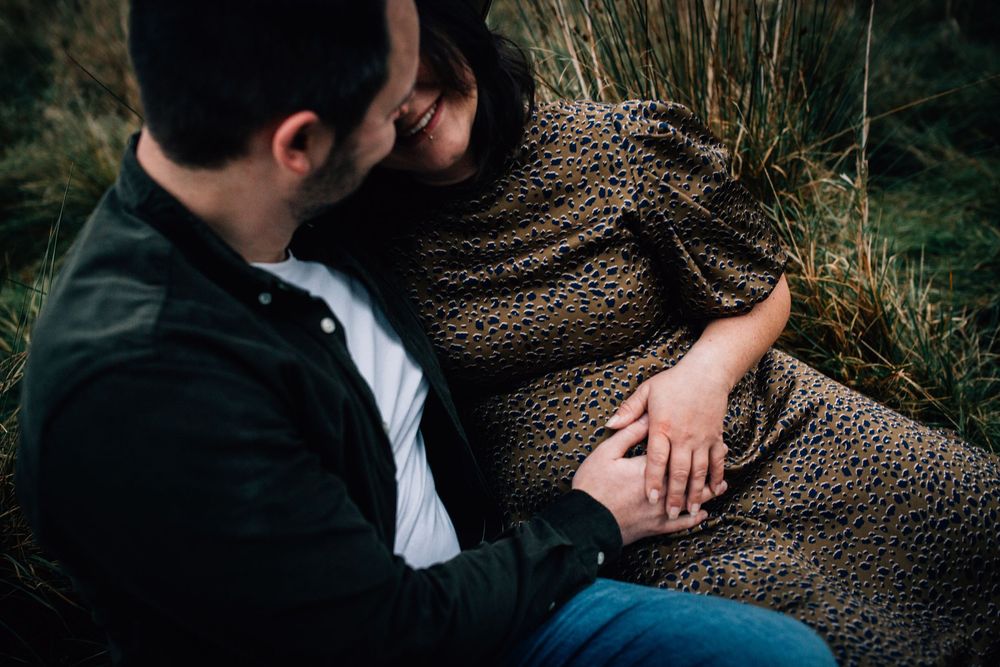 Couple sitting outdoors holding hands, woman in leopard print dress leaning on man in green jacket during maternity photo session.
