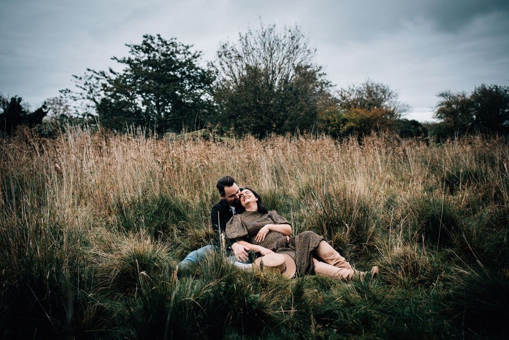 Couple sitting together in tall golden grass field, embracing romantically under cloudy autumn sky.