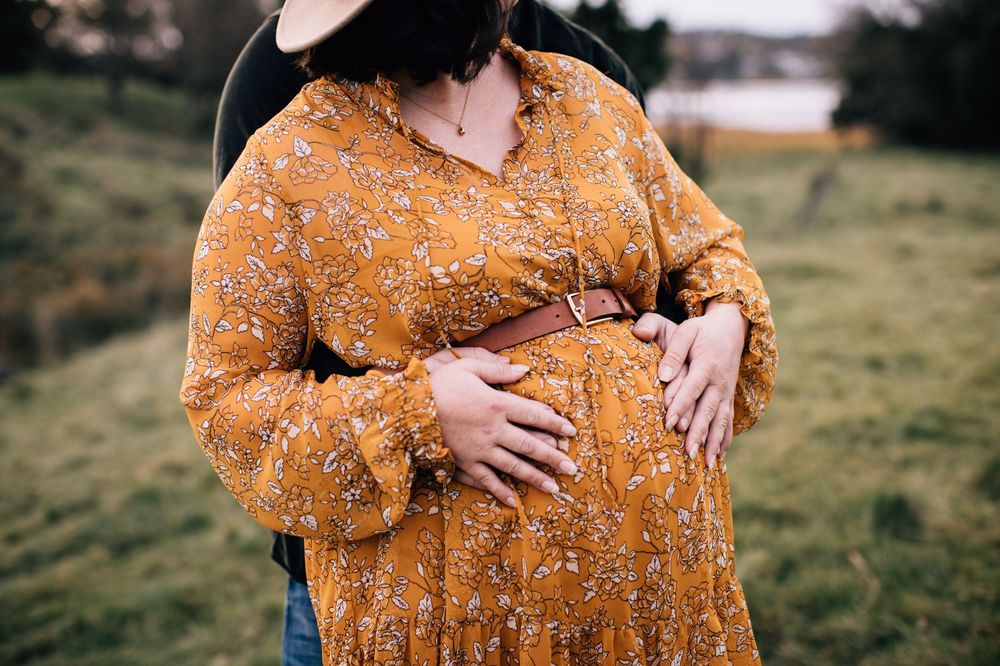 Pregnant woman in mustard floral dress holding baby bump outdoors in green field.