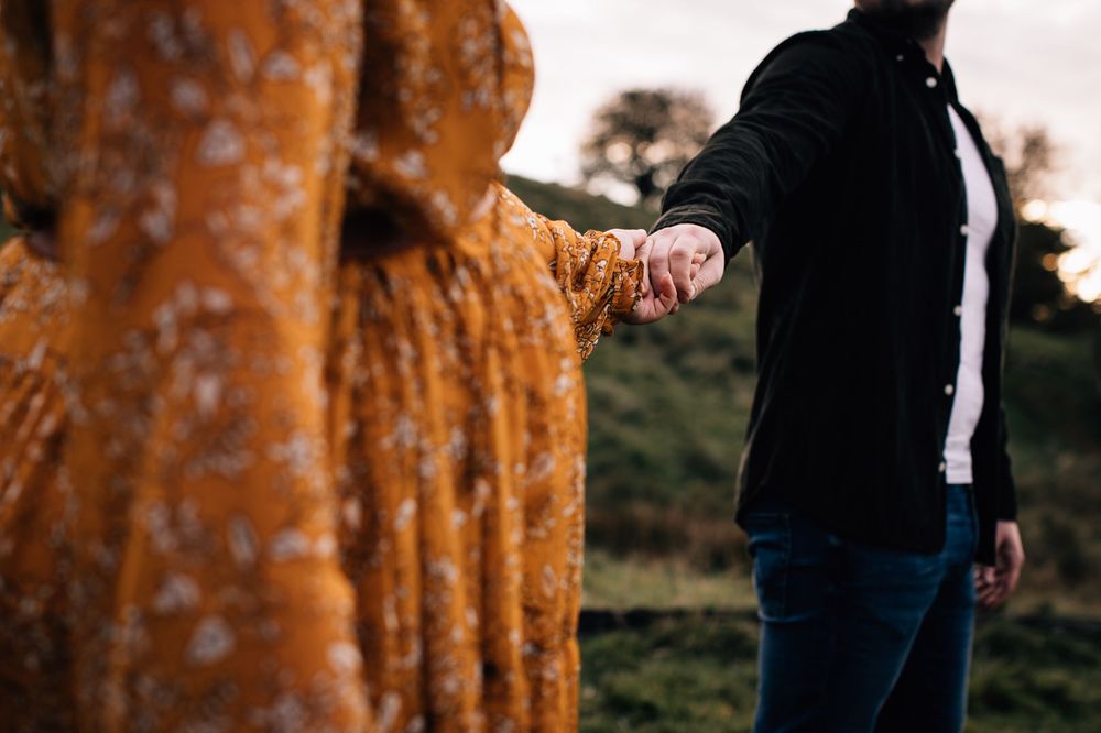 Couple holding hands outdoors at sunset, woman in floral orange dress and man in black jacket with blue jeans.
