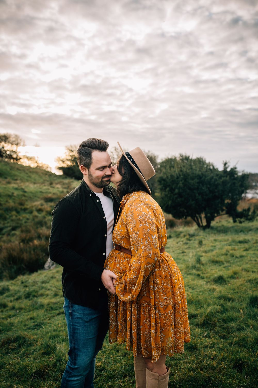 A couple shares a romantic moment in a green field, woman in yellow floral dress and hat kissing man in black shirt.