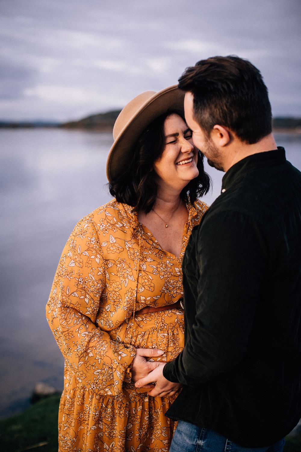Couple sharing a tender moment by a lake, woman in golden floral dress and wide-brim hat smiling joyfully.