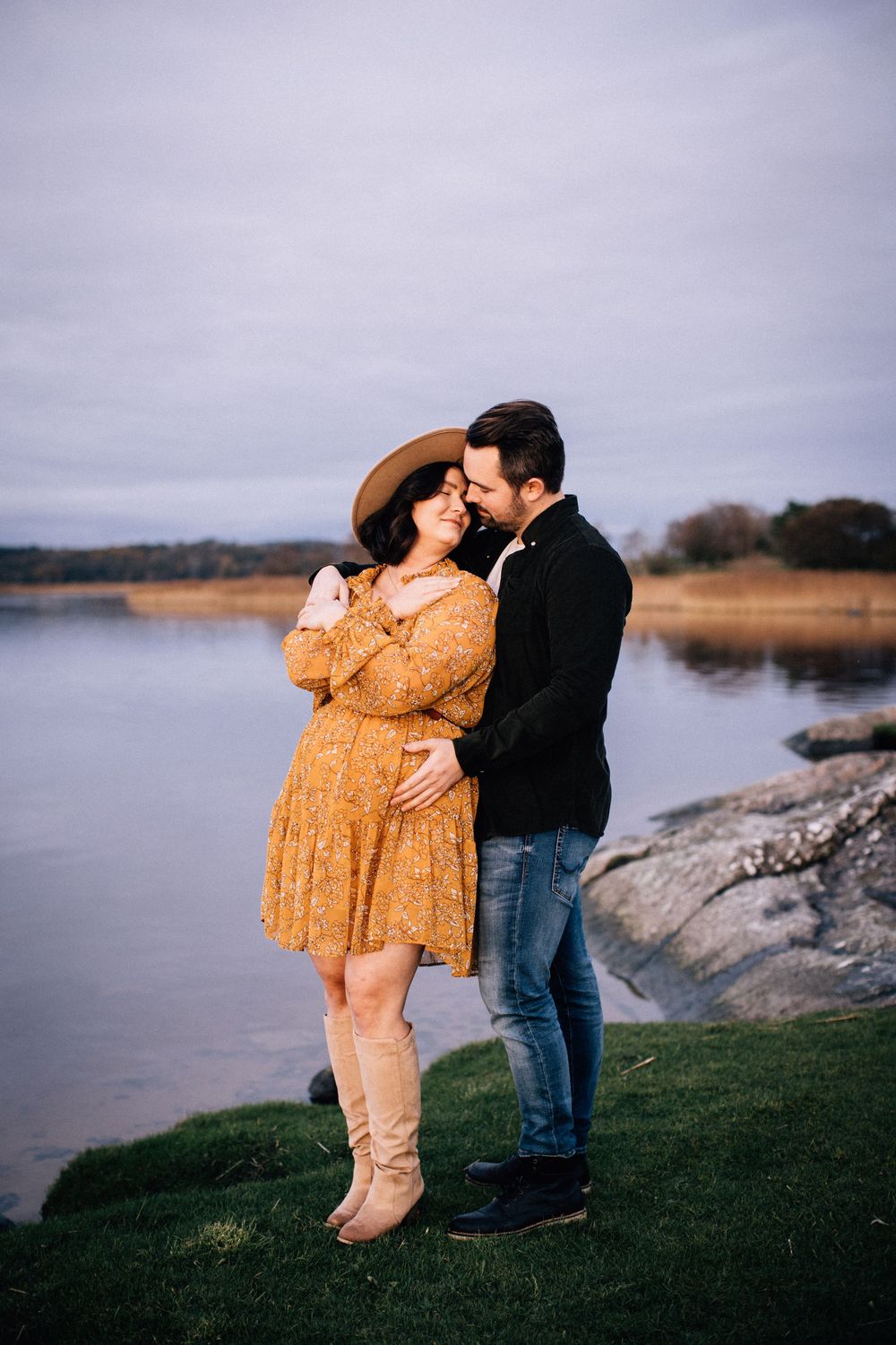 Couple embracing near a lake, pregnant woman in orange floral dress and hat, man in black jacket and jeans.