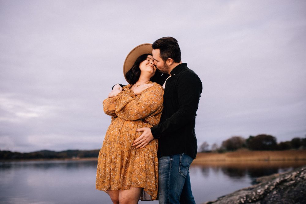 Couple sharing a romantic kiss near a lake, woman in floral dress and hat, man in black jacket, cloudy sky.