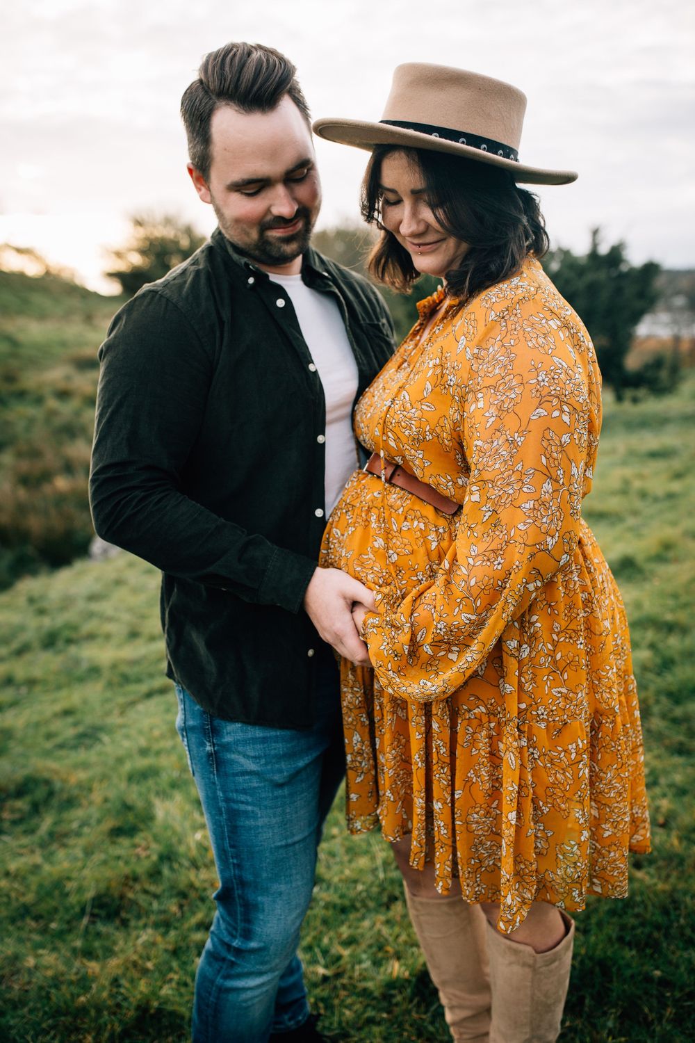 A couple shares a tender moment outdoors, with the woman in a golden floral dress and hat cradling her baby bump.
