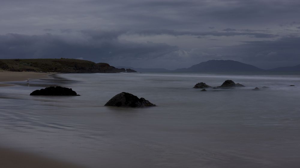 Moody coastal beach scene with dark rocks emerging from calm waters under overcast skies with distant mountains.