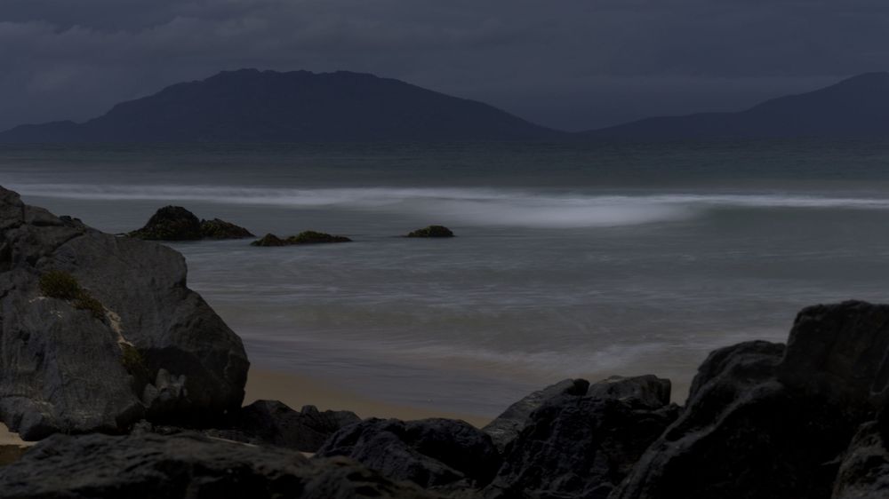 Rocky shoreline with misty ocean waves and dark mountain silhouettes under a stormy sky.
