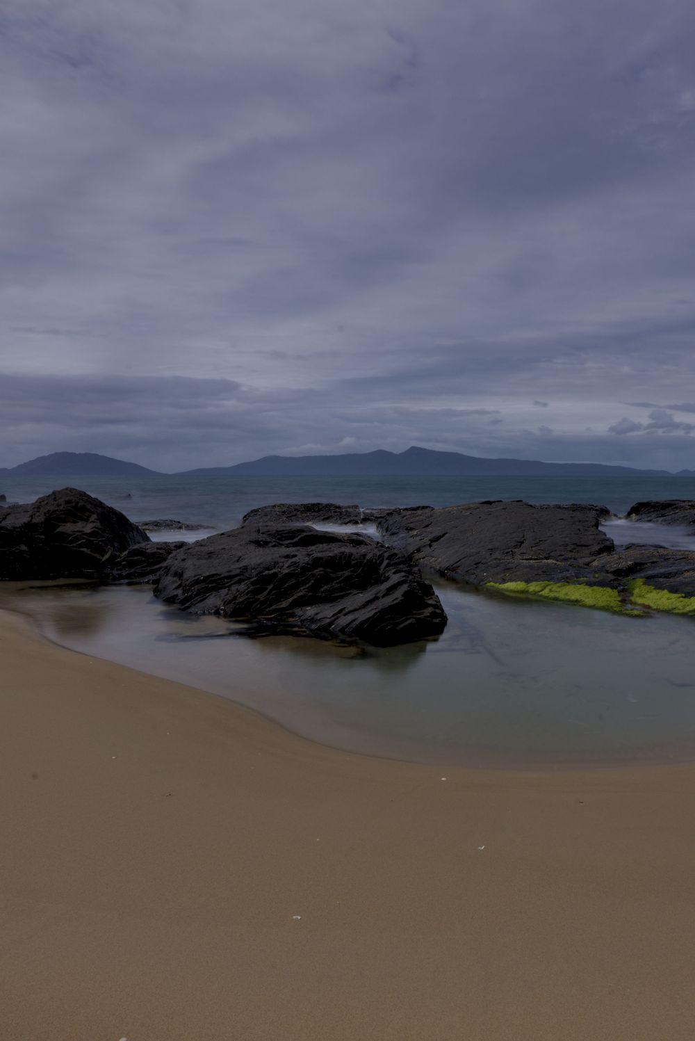 Rocky beach shoreline with dark boulders, green algae, calm tidal pools, sandy shore, and distant mountains under dramatic cloudy sky.