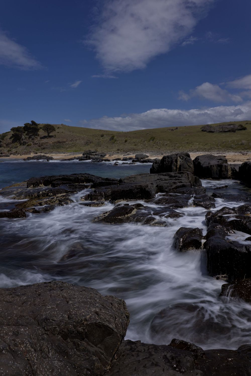 Rocky coastal shoreline with crashing waves, green hills, and blue sky with white clouds in a scenic seascape.