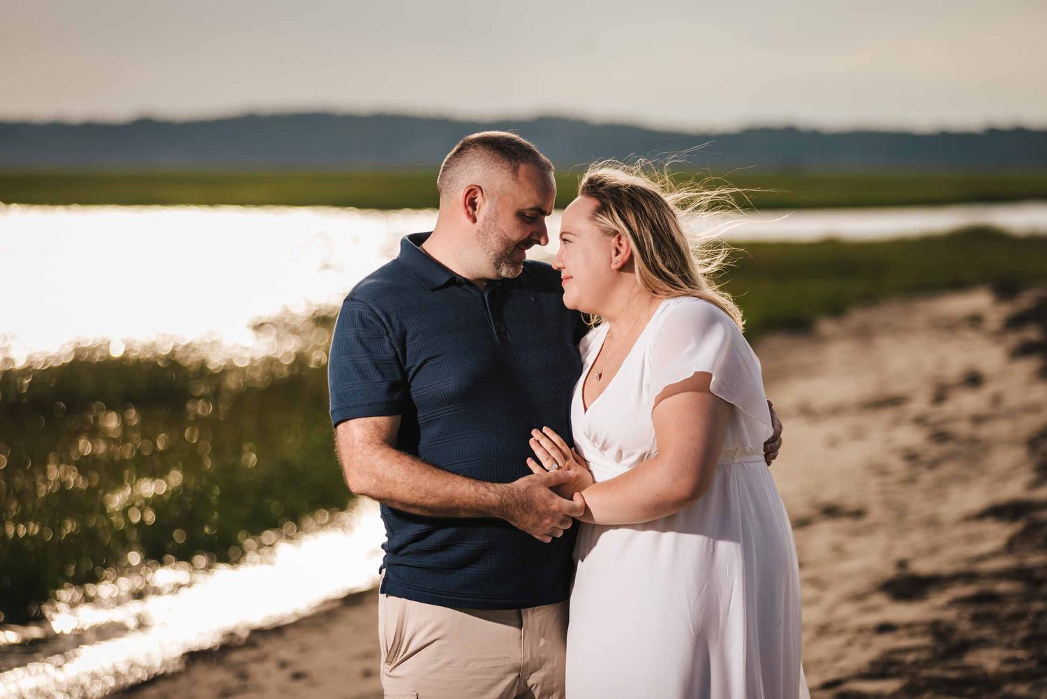 Engaged couple embracing on the sand at Duxbury Beach, Massachusetts, with backlit marsh grass and water behind them.