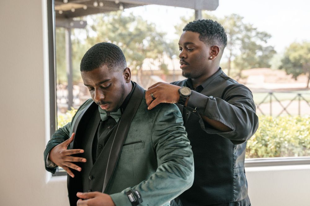 Two well-dressed men in suits, one adjusting the other's jacket near a bright window.