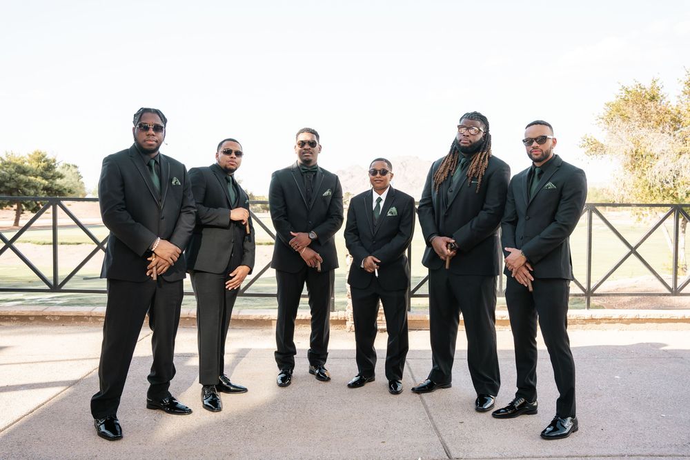 Six men in matching black suits posing together outdoors at a formal event, likely a wedding party.