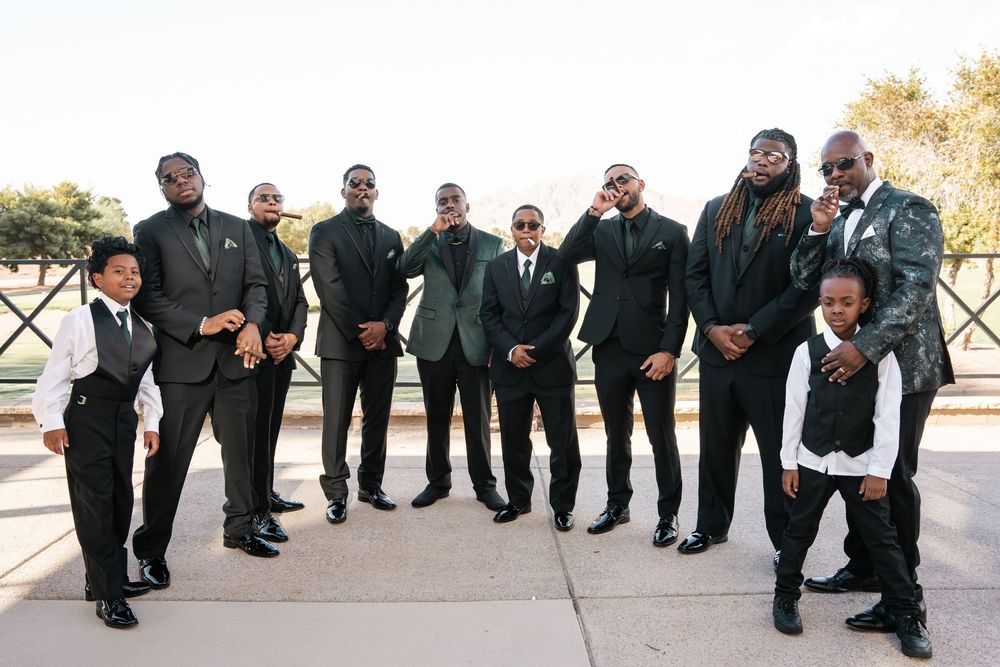 Groom in green tuxedo poses with groomsmen in black suits outdoors at elegant wedding venue.