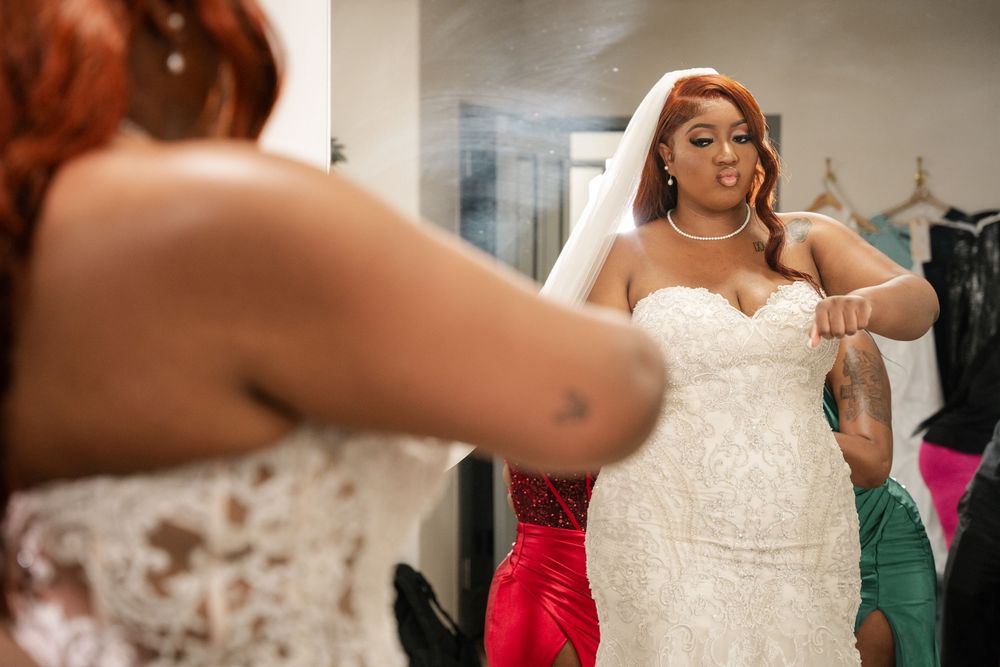 Bride in white lace wedding gown and veil being assisted by bridesmaid in red dress in a bridal suite.