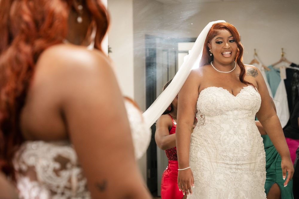 A plus-size bride in a beaded strapless wedding gown and veil smiles at her reflection in a mirror.
